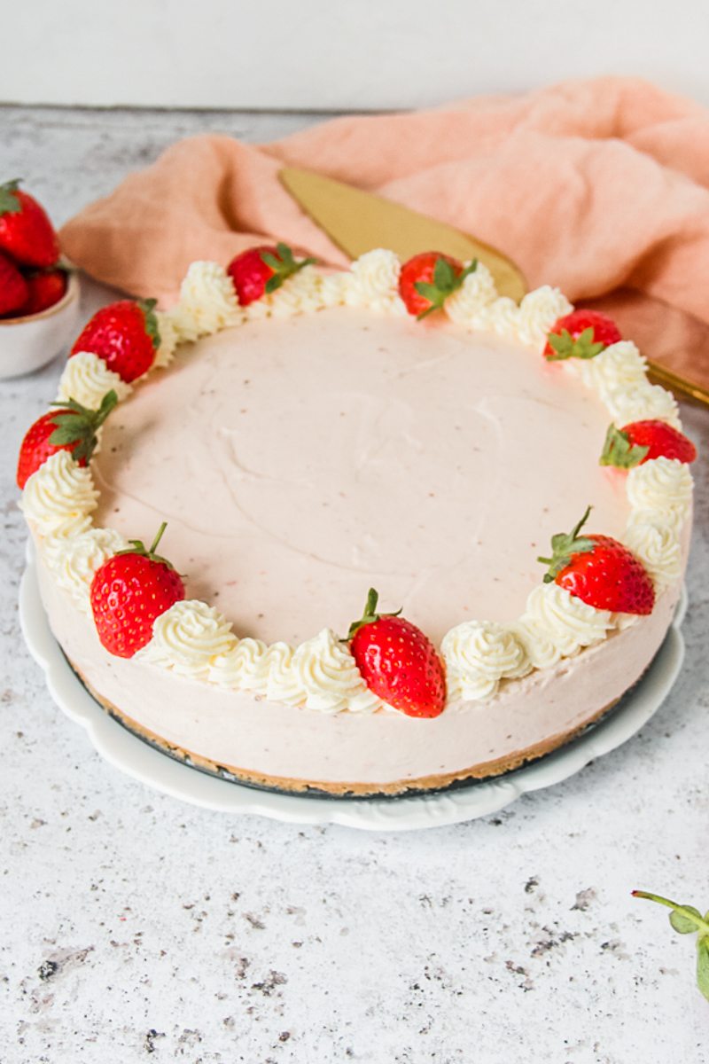 A no bake strawberry cheesecake topped with a whipped cream border and strawberries sits on a white plate on a light gray surface.