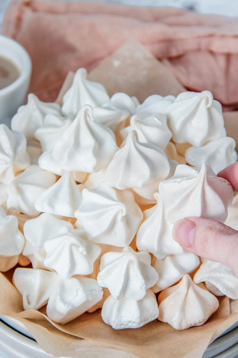 A Suspiros Cookie is picked up from a bunch sitting on a stack of plates and parchment paper on a light gray surface with a coral cloth behind.