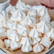 Suspiros - Latin Meringues Cookies sit on a stack of plates and parchment paper on a light gray surface with a coral cloth behind.