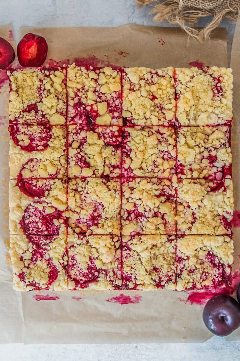 Svestkový tác - Czech Plum Cake sits on baking paper on a gray surface with 16 slices into the square cake.