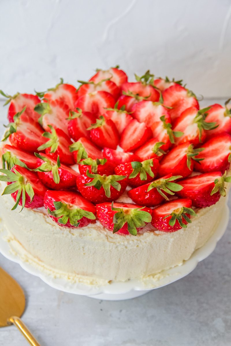 Halved strawberries decorate the top of a three layer sponge cake decorated with whipped cream on a white fluted cake stand on a gray surface.