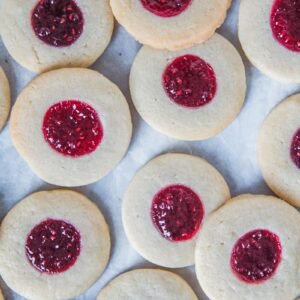 A close up of raspberry jam Swedish thumbprint cookies laying unevenly on a parchment paper lined gray surface.