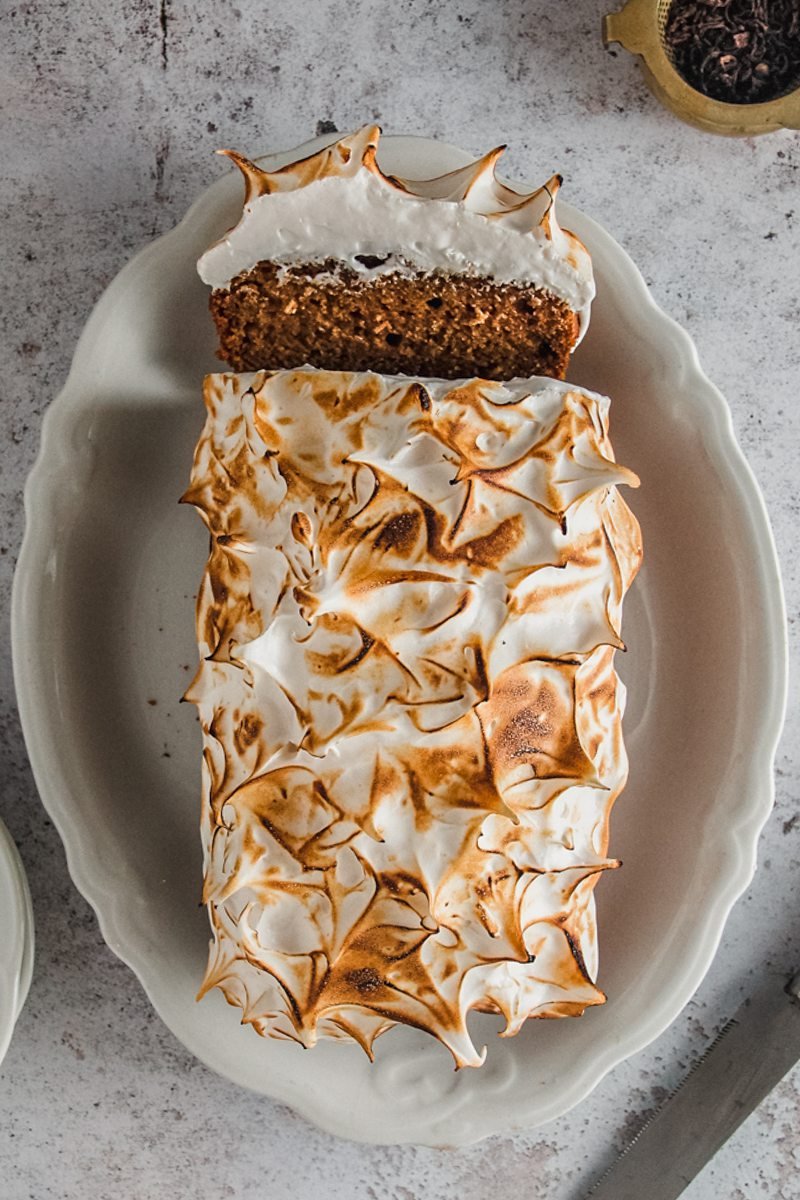 A sweet potato loaf cake topped with a homemade caramelized marshmallow topping sits on an oval white ceramic plate on a light gray surface with one slice resting against the edge of the plate.