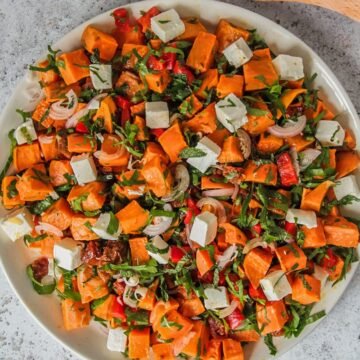 A sweet potato salad sits on a round white ceramic plate on a gray surface with wooden salad serves on the side.