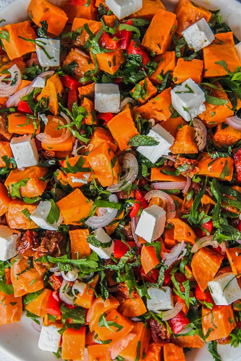 A close up of sweet potato salad with cubes of feta and sweet potato scattered among other herbs and vegetables.