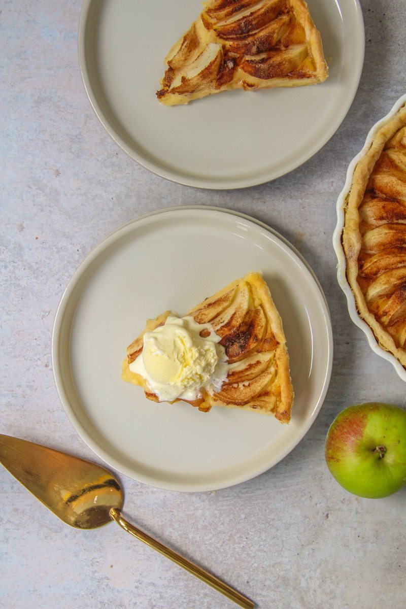 A slice of Portuguese Apple Tart sits on a rimmed ceramic plate with a scoop of vanilla ice cream on top. A second slice sits nearby without alongside the remaining tart, gold server and apple on a gray surface.