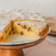 A slice of Teisan Lap - Welsh Raisin Cake sits on a white fluted plate on a wooden cake stand on a light pink surface.