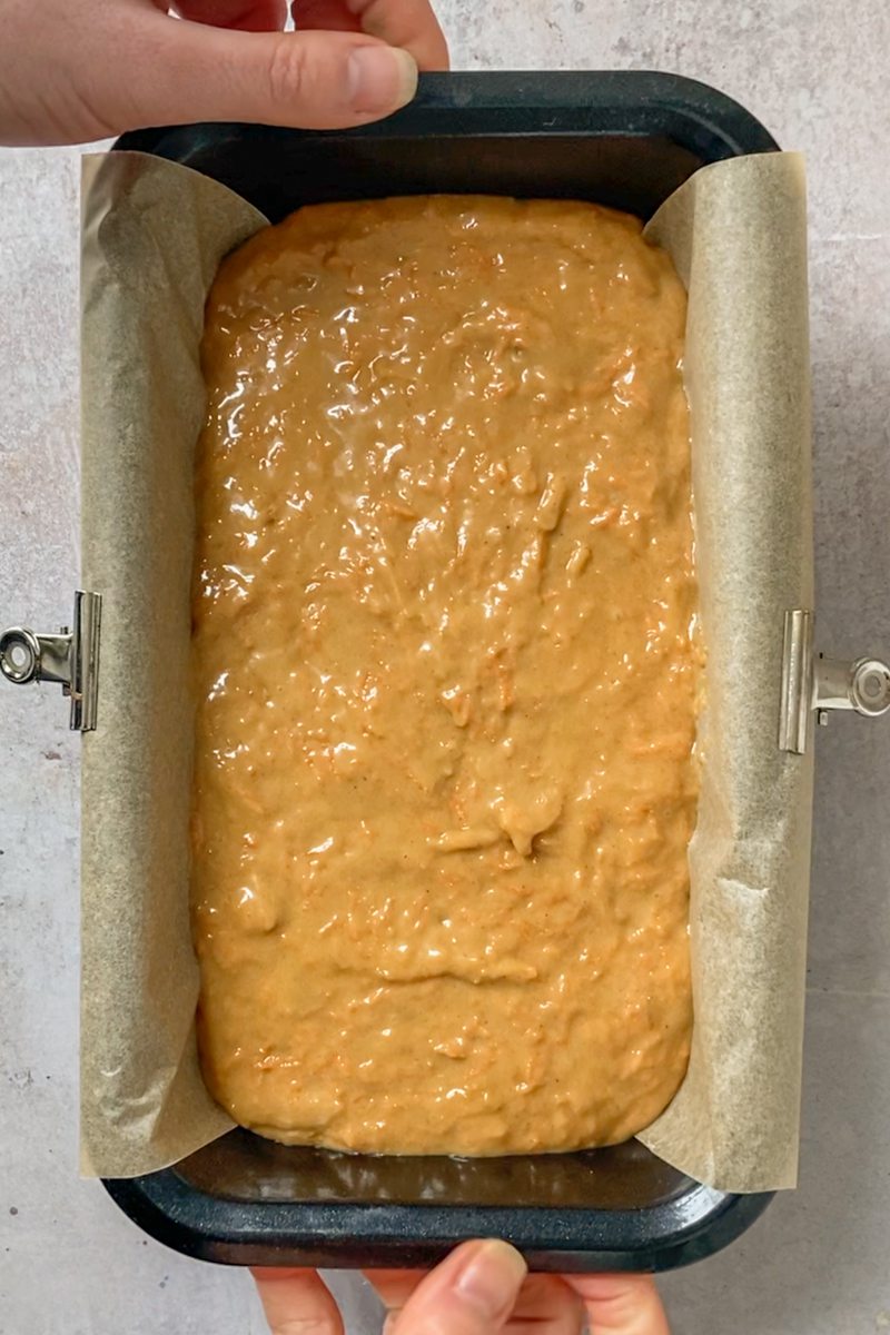 Unbaked carrot cake loaf sits in a lined metal black loaf tin on a gray surface.