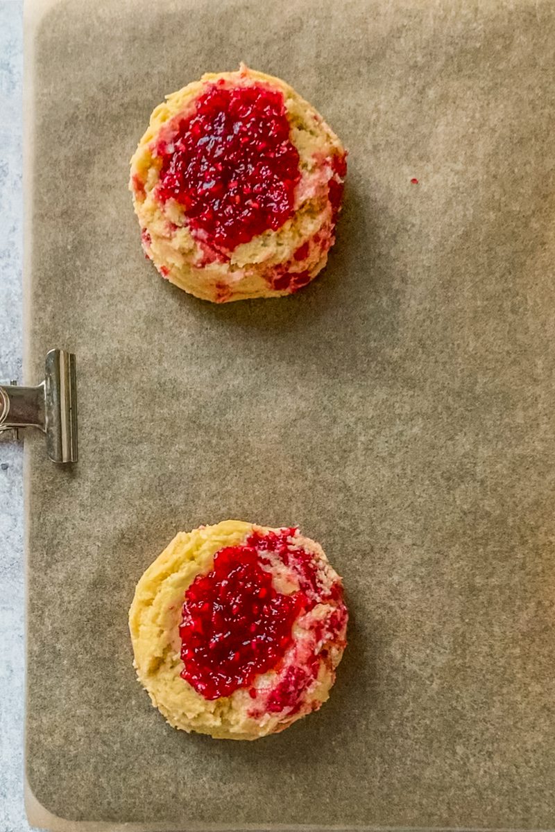 Two unbaked white chocolate raspberry cookies sit on a lined baking tray.