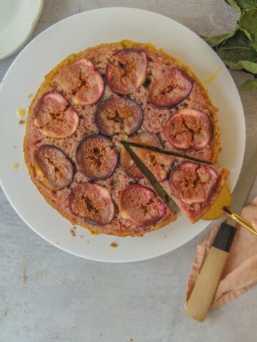 A slice of upside down fig cake sits on a cake server with the remaining cake surrounding it on a round white ceramic cake above a gray surface