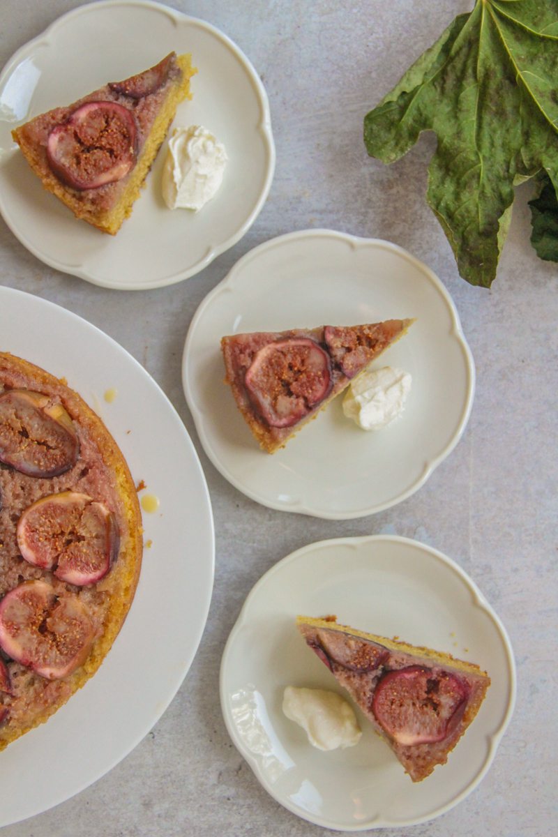 Slices of upside down Fig cake sit on scalloped white ceramic plates on a gray surface with the remaining cake beside and some dried fig leaves nearby.