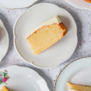 Slices of vanilla cake with cream cheese frosting sit on individual slices on a light gray surface.