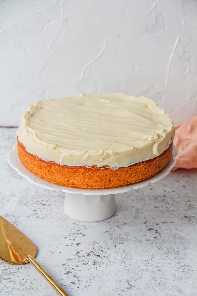 A vanilla cake with cream cheese frosting sits on a white ceramic cake stand on a light gray surface.