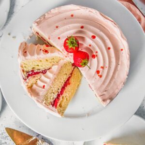 Slices of vanilla strawberry cake with a roasted strawberry center and strawberry Swiss meringue buttercream sits on a white ceramic plate on a light gray surface with a coral colored cloth beside.