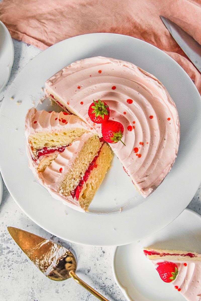 Slices of vanilla strawberry cake with a roasted strawberry center and strawberry Swiss meringue buttercream sits on a white ceramic plate on a light gray surface with a coral colored cloth beside.