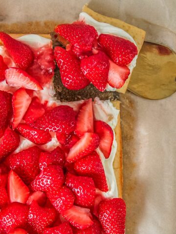A slice of small vanilla sheet cake topped with whipped cream and strawberries sits on a cake server beside the remaining cake on parchment paper.