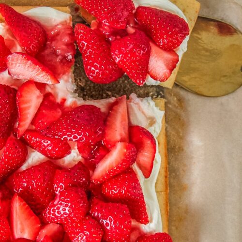 A slice of small vanilla sheet cake topped with whipped cream and strawberries sits on a cake server beside the remaining cake on parchment paper.