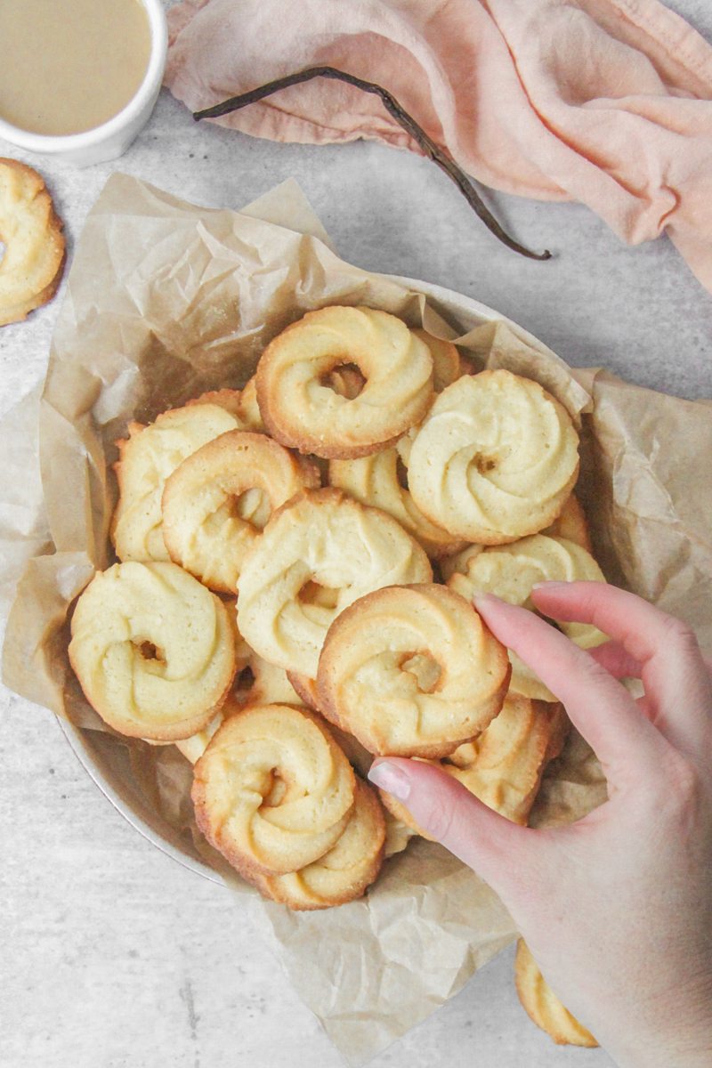 A Vanillekranse - Danish butter cookie is held above other cookies sitting in a deep ceramic plate lined with parchment paper on a gray surface.