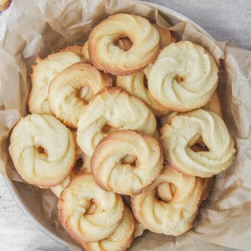 Vanillekranse - Danish butter cookies sit in a deep ceramic plate lined with parchment paper on a gray surface with tea beside.