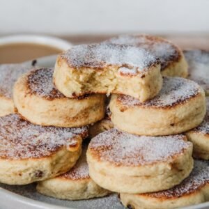 A stack of Welsh cakes sit on a rimmed ceramic plate with the top cake having a bite out of it.