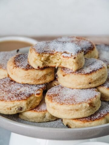 A stack of Welsh cakes sit on a rimmed ceramic plate with the top cake having a bite out of it.