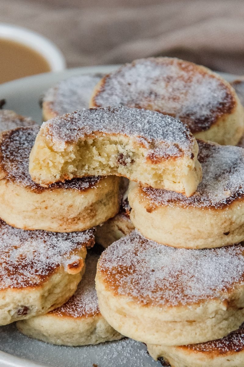 A stack of Welsh cakes sit on a rimmed ceramic plate with the top cake having a bite out of it.