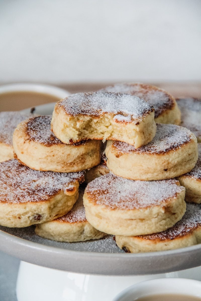 A stack of Welsh cakes sit on a rimmed ceramic plate with the top cake having a bite out of it.