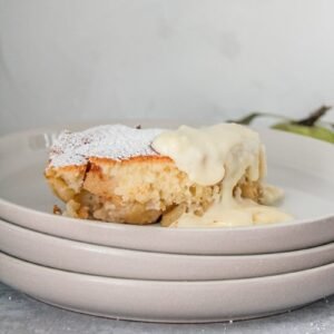 A serving of Welsh Eves pudding sits on a stack of rimmed grey ceramic plates with a serving of custard on the right hand corner.