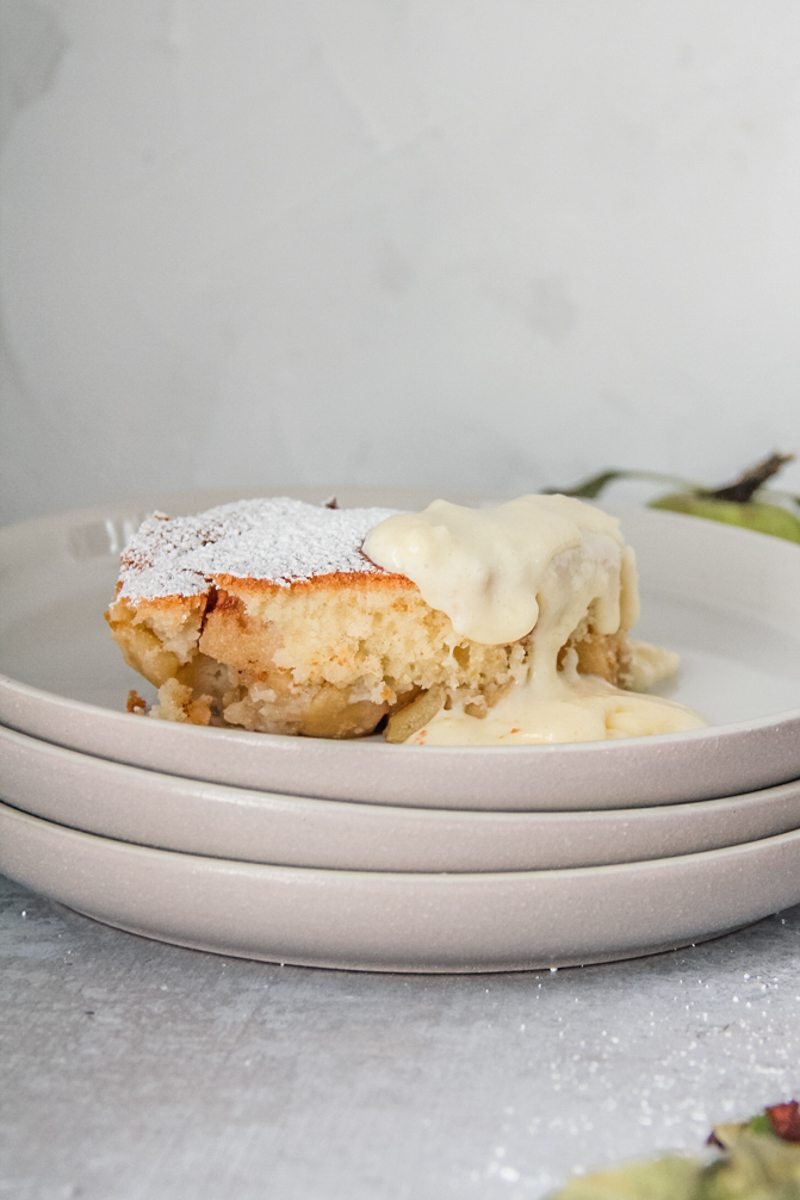 A serving of Welsh Eves pudding sits on a stack of rimmed grey ceramic plates with a serving of custard on the right hand corner.