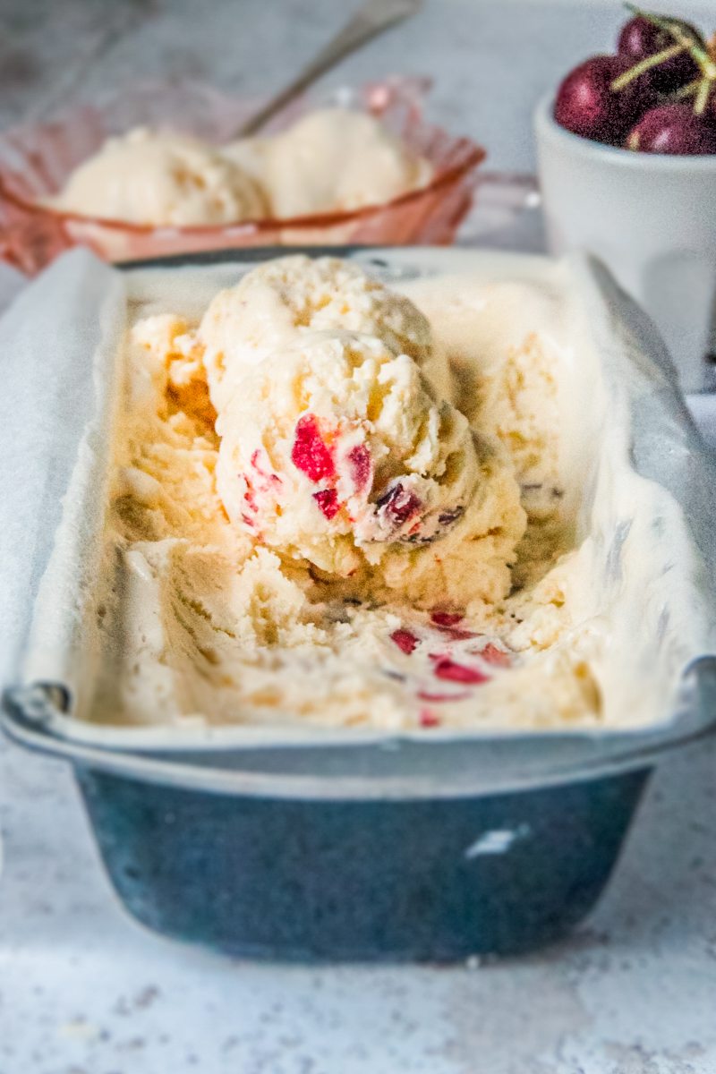Scoops of cherry white chocolate no churn ice cream sit on top of a lined rectangle metal tin on a light gray surface.