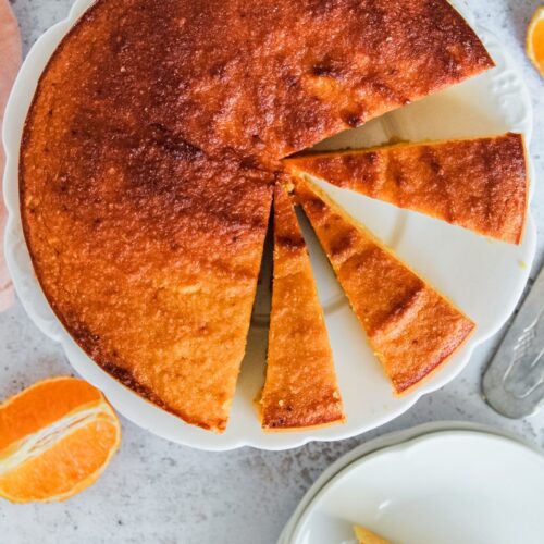 Whole orange cake sits on a white plate with a few slices cut out and a slice on an individual plate beside on a gray surface.