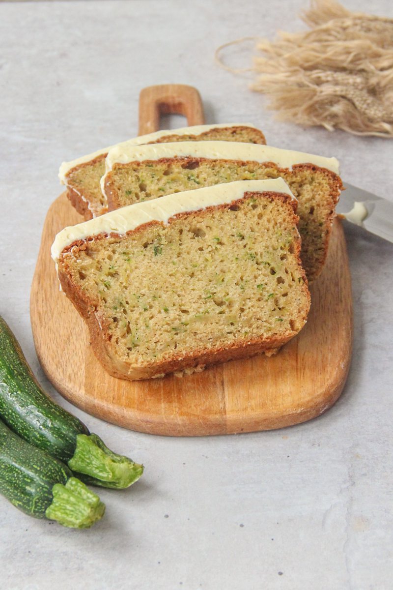 Slices of zucchini loaf cake with cream cheese frosting rest on a wooden board on a gray surface.