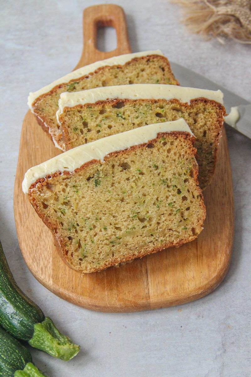 Slices of zucchini loaf cake with cream cheese frosting sit on a wooden board on a gray surface.