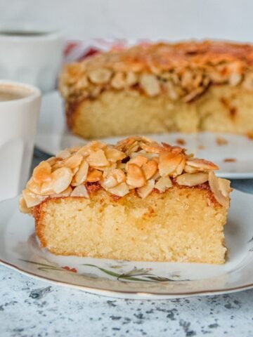 A slice of Swedish Almond Cake sits on a ceramic plate on a light gray surface.