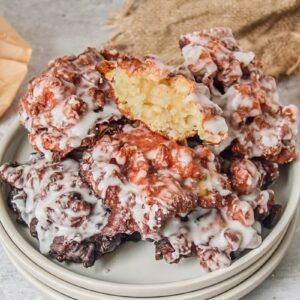 Amish apple fritters sit stacked on a stack of rimmed ceramic plates on a light gray surface with the top fritter showing the light apple interior visible.