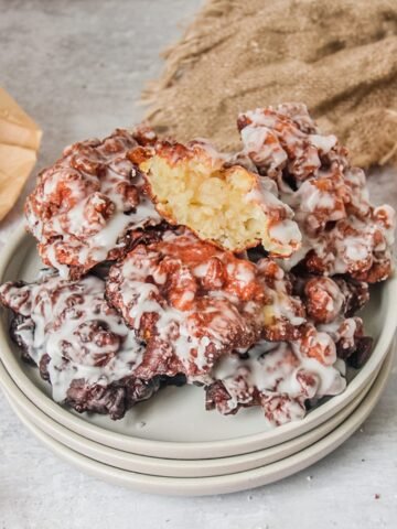 Amish apple fritters sit stacked on a stack of rimmed ceramic plates on a light gray surface with the top fritter showing the light apple interior visible.