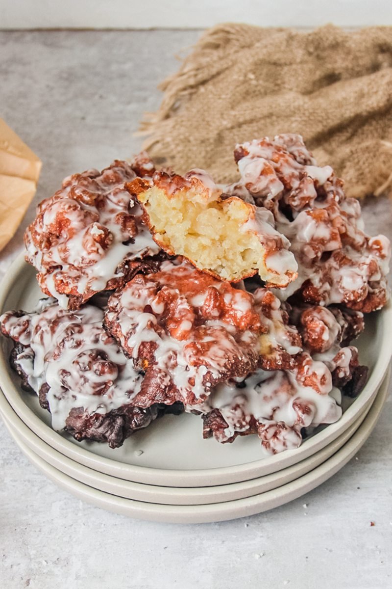 Amish apple fritters sit stacked on a stack of rimmed ceramic plates on a light gray surface with the top fritter showing the light apple interior visible.