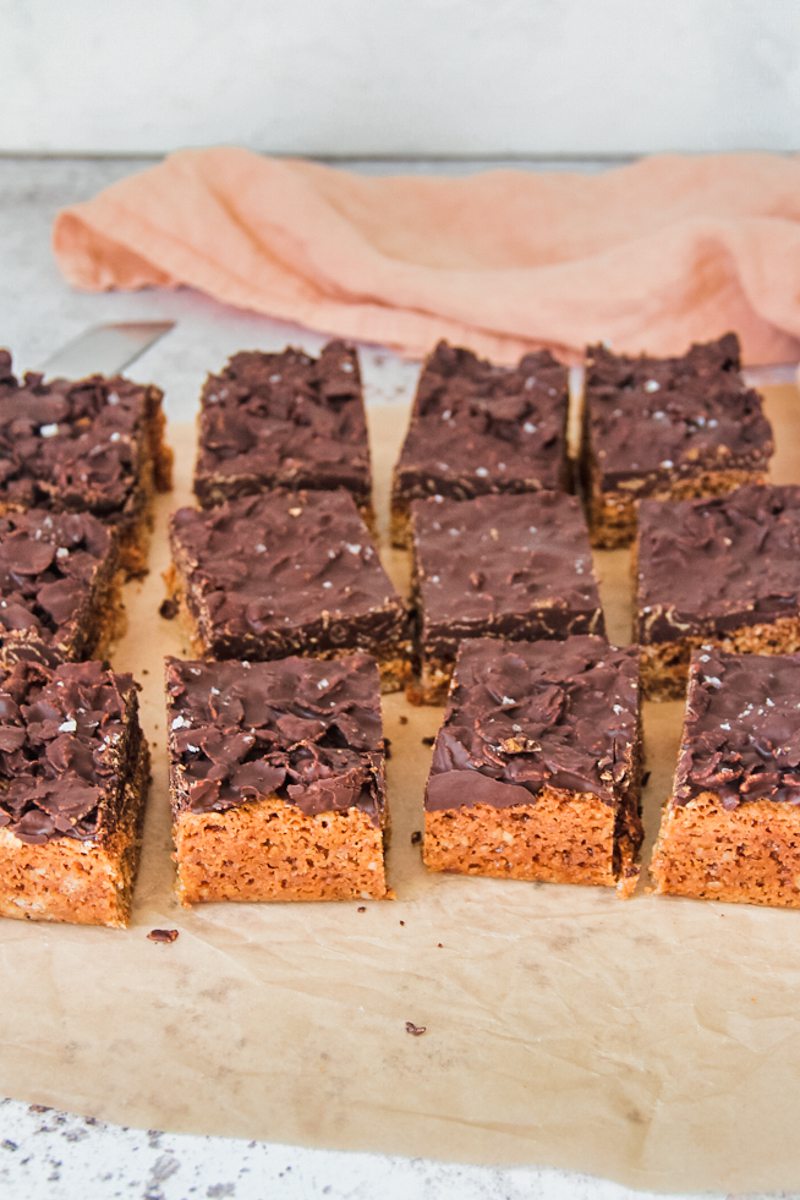 Anzac biscuit bars sit on parchment paper on a light gray surface with a coral cloth behind.