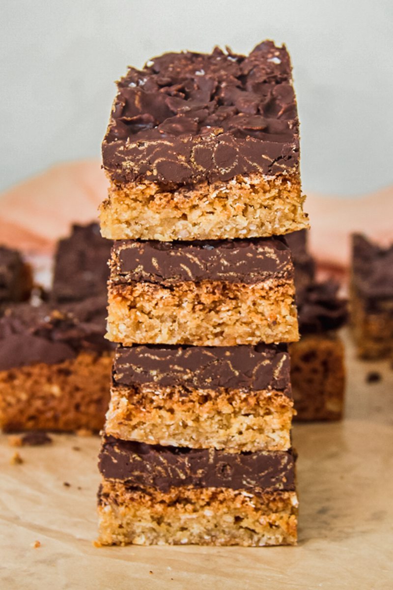 A stack of Anzac biscuit bars sit on top of each other on a parchment paper with remaining biscuit bars behind.