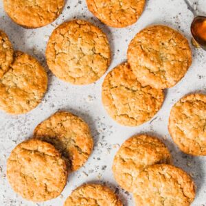 Anzac biscuits lay on a light gray surface with oats and a spoonful of golden syrup beside.