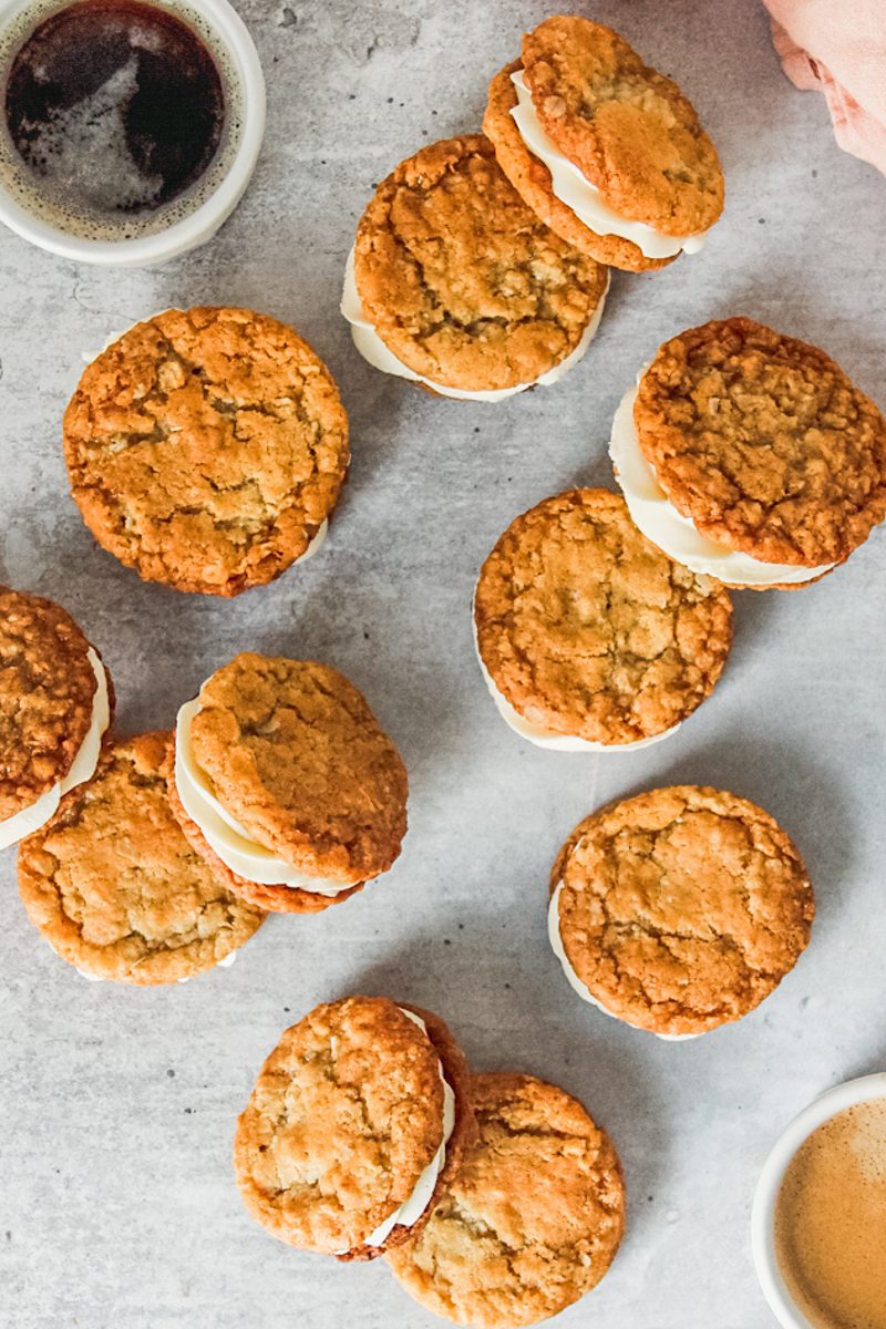 Anzac sandwich biscuits with a white chocolate ganache filling lay on a gray surface beside two cups of espresso.