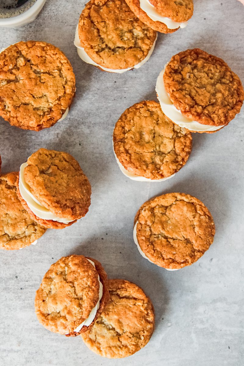 Anzac sandwich biscuits with a white chocolate ganache filling lay on a gray surface beside a cup of espresso.