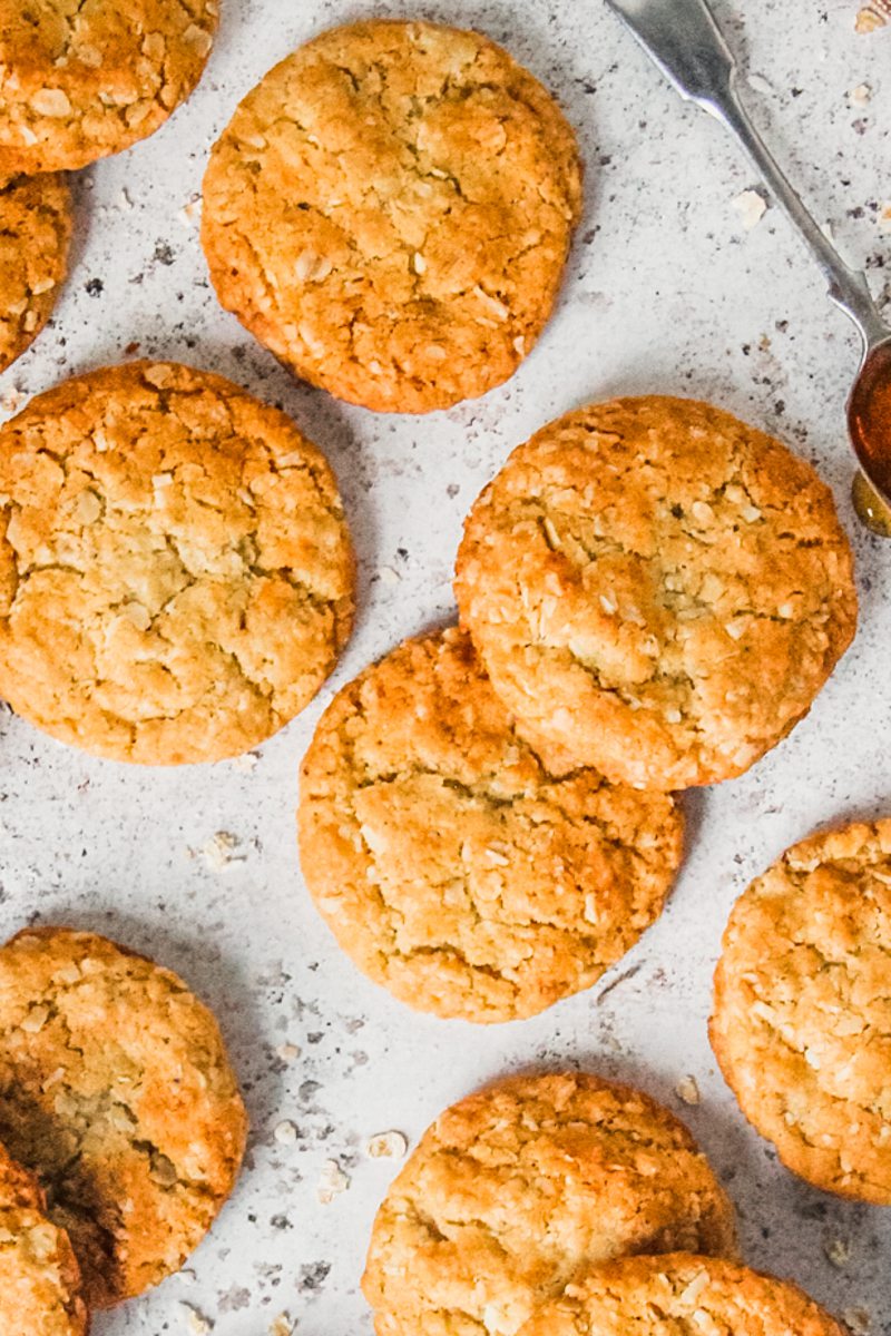 Anzac biscuits lay on a light gray surface with a spoonful of golden syrup beside.