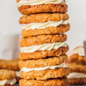 A stack of anzac sandwich biscuits with a white chocolate ganache center sit on a gray surface with remaining cookies behind.