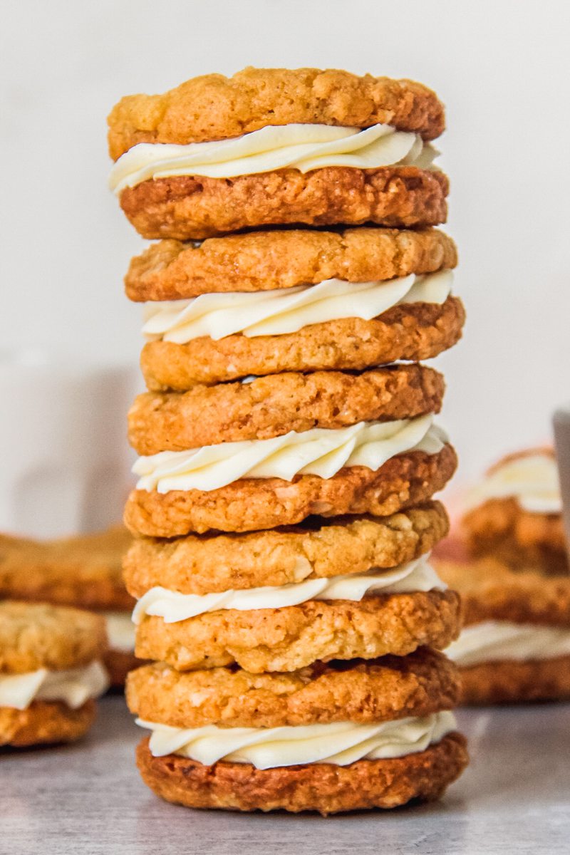 A stack of anzac sandwich biscuits with a white chocolate ganache center sit on a gray surface with remaining cookies behind.