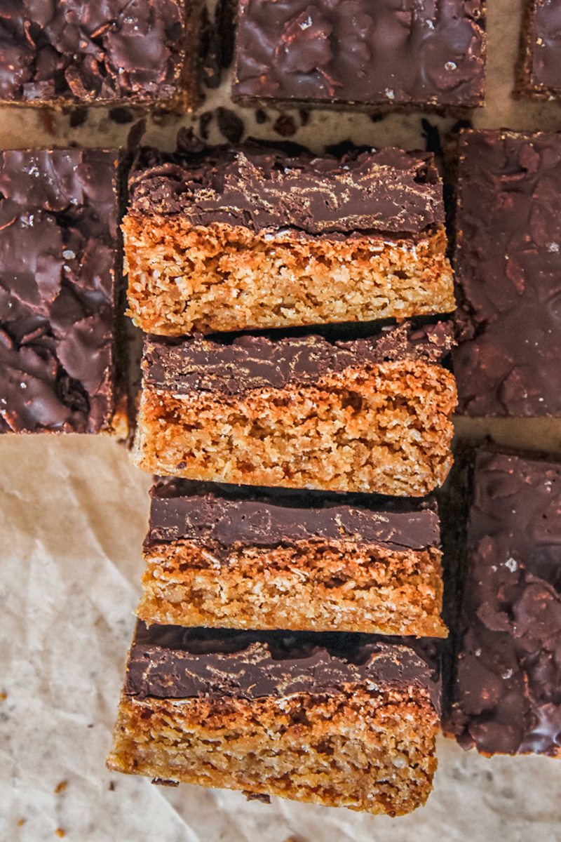 Anzac biscuit bars lay stacked on top of each other on a brown parchment paper.