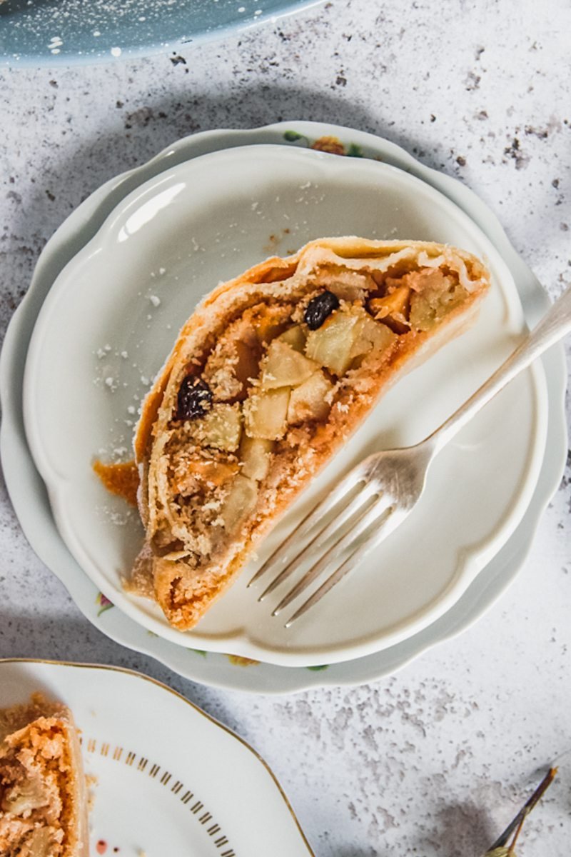 A slice of apple strudel sits on a stack of white plates on a light gray surface with a fork on the side.