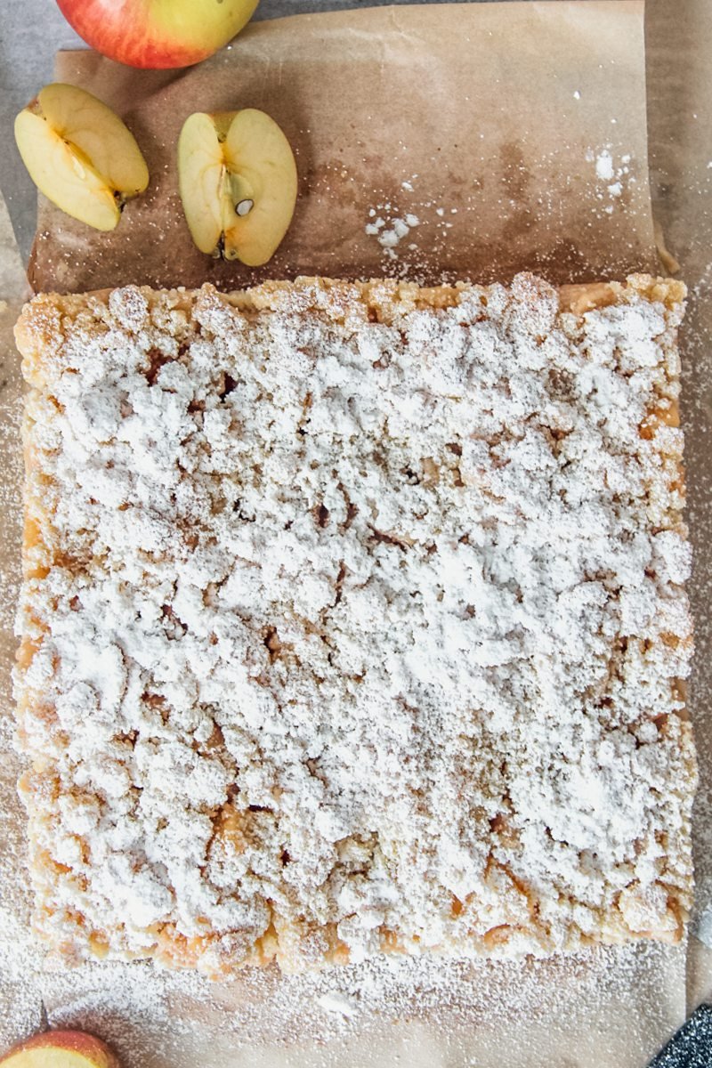 Apple crumble slice coated in powdered sugar sits on brown parchment paper with apple slices.