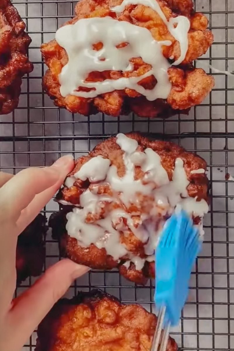 A glaze is brushed over apple fritters on a cooling tray on a gray surface.