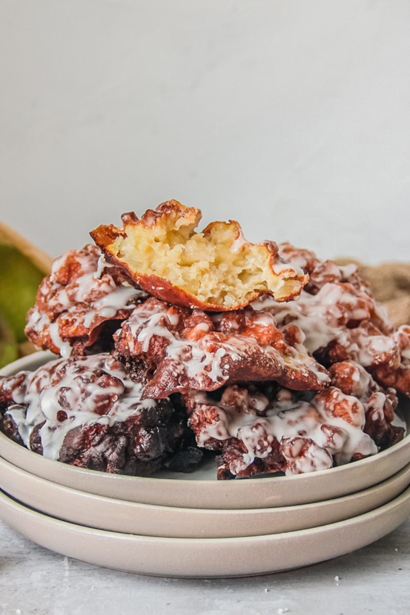 Amish apple fritters sit stacked on a stack of rimmed ceramic plates on a light gray surface with the top fritter broken in half to show the light apple interior.